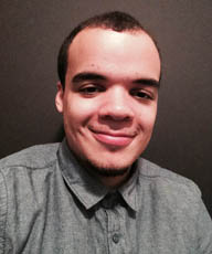 Brandon Williams, a light skinned man with dark brown hair, and a light denim blue shirt. The background is a black wall. Brandon has a closed smile.